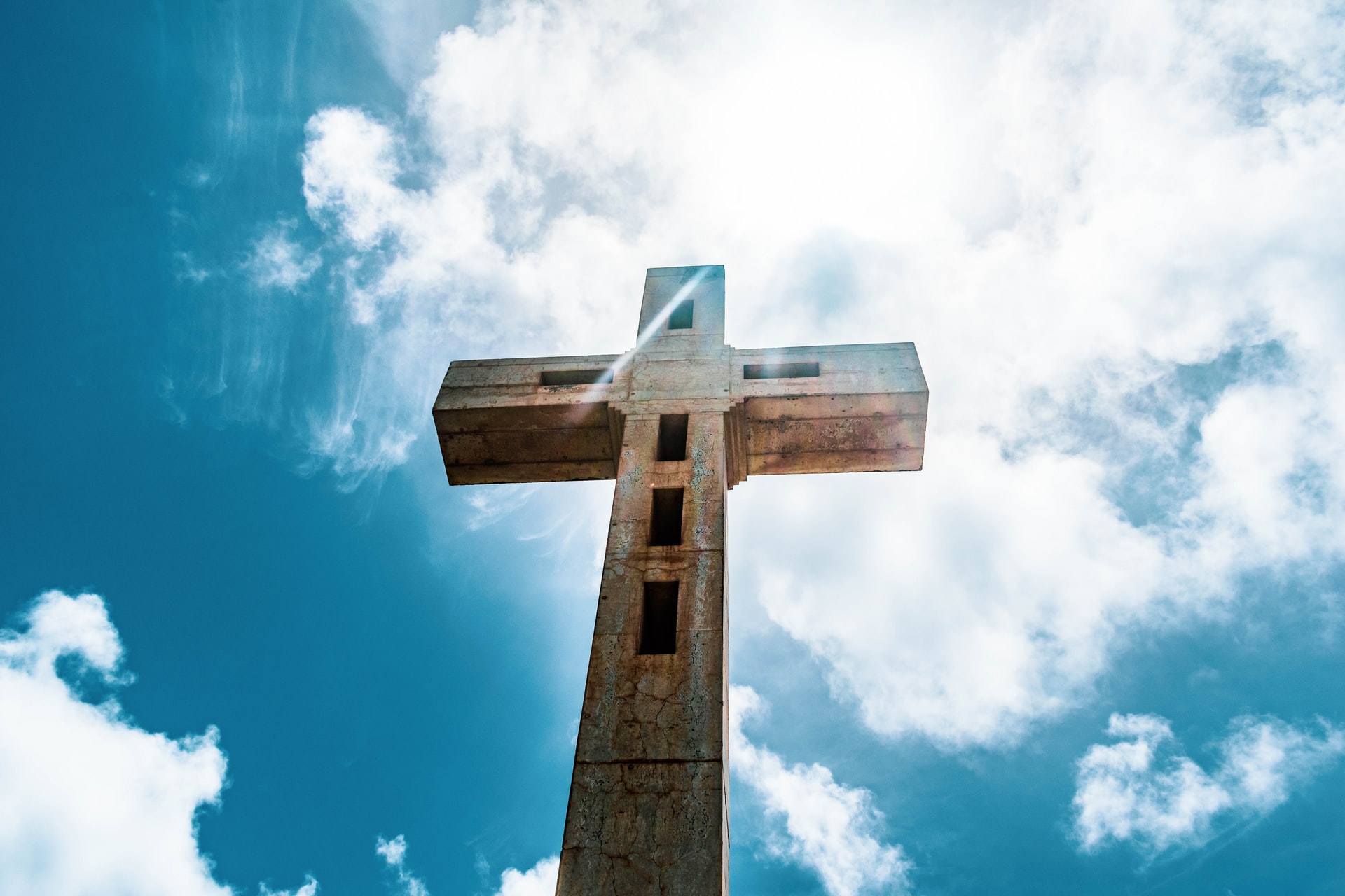 Looking up at a large cross against a blue sky with many white clouds and bright white sunlight