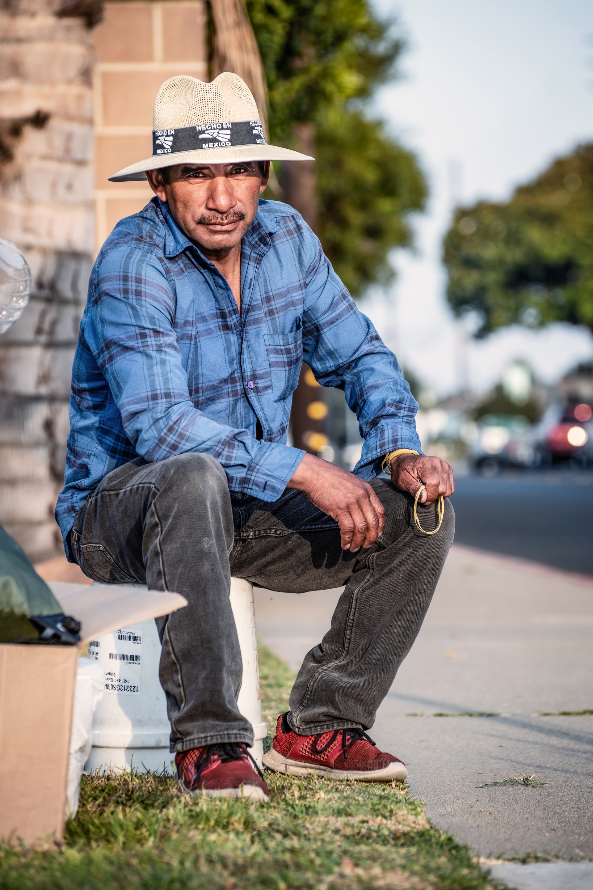 An older Hispanic man wearing a white hat, blue long sleeved shirt, black pants, and red shoes seated on an overturned white bucket alongside a street while facing the camera