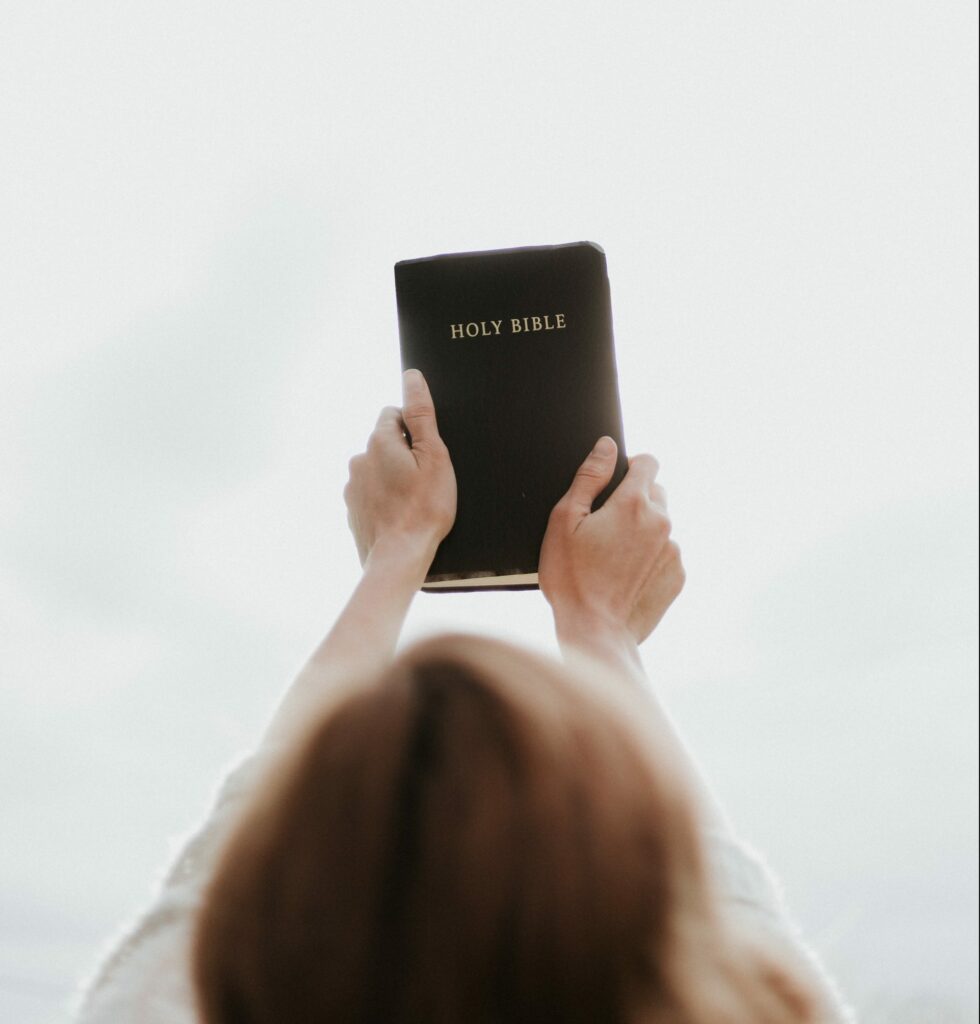 Looking across the back of the head of young Caucasian woman with brown hair holding a black covered Bible at arms length against a white background, showing her faith.