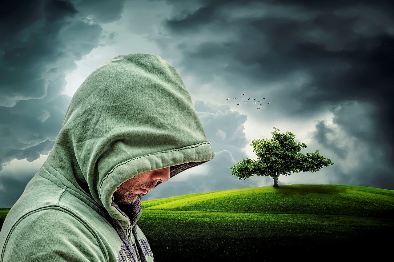 A graphical image of a man in a light emerald green hooded sweat shirt looking down while hidden in hood with a clouded sky, a partially lit hill, and tree in the background