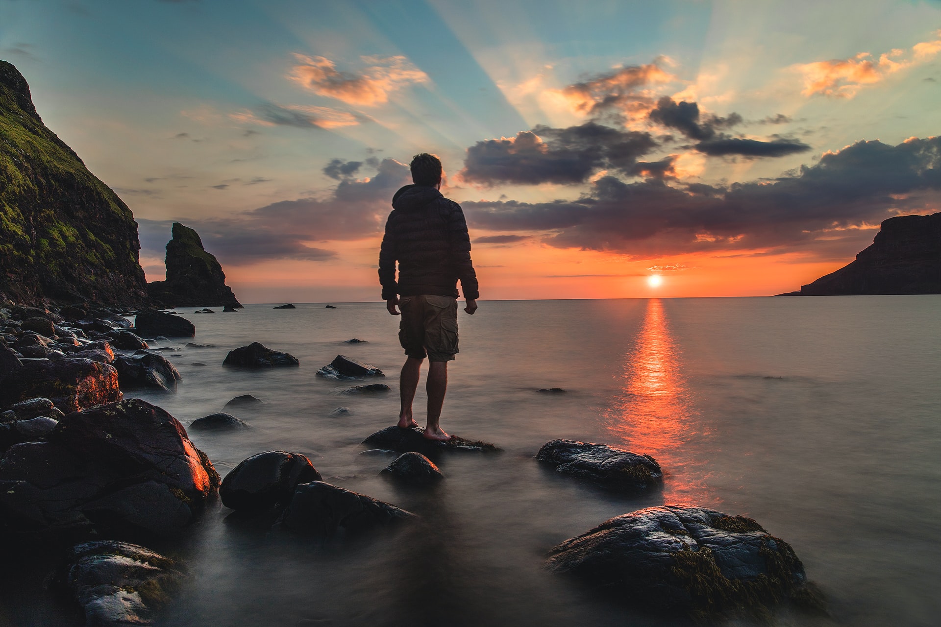 A partial silhouette of the backside of a man wearing a hooded jacket and shorts standing on one of a series of rocks near the rocky shore of a large body of water while looking at a sunset with a small mountain and clouds in the distance
