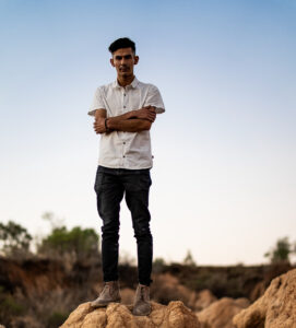 A photo of a young Hispanic man standing on a cluster of desert rocks with brush and a blue sky in the background