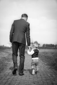 Black and white photo of man and boy in suits holding hands while walking away down a brick walkway