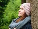 A photo of a side headshot of a young Caucasian woman with brown hair wearing a lavender crocheted hat, a medium light blue scarf, and a dark blue jacket, looking upward while leaning against a tree with green shrubbery in the background