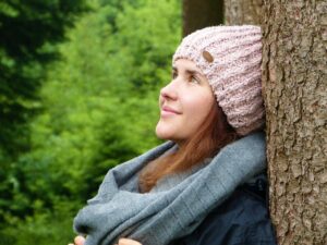 A photo of a side headshot of a young Caucasian woman with brown hair wearing a lavender crocheted hat, a medium light blue scarf, and a dark blue jacket, looking upward while leaning against a tree with green shrubbery in the background