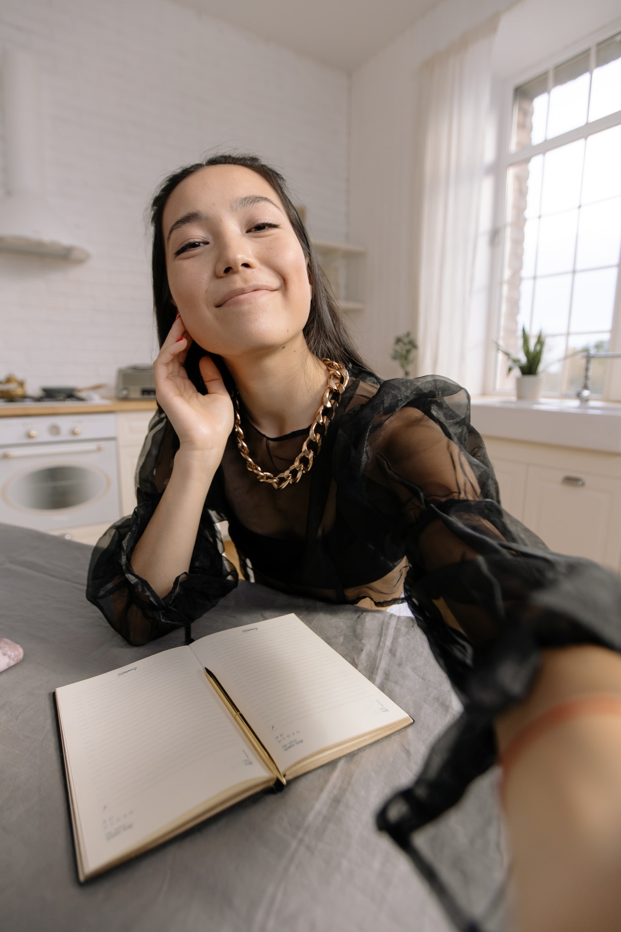 A selfie photo of a young Asian woman wearing a black blouse and a large gold necklace resting her right elbow on a table while holding her hand near her neck, an open notebook rests on the table in a large, all white modern kitchen in an attempt to satisfy herself.