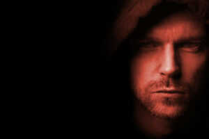 A lightly red tinted photo of the headshot of a Caucasian man with a mustache and light beard positioned to the far right wearing a brown hood and displayed with a serious expression on his face