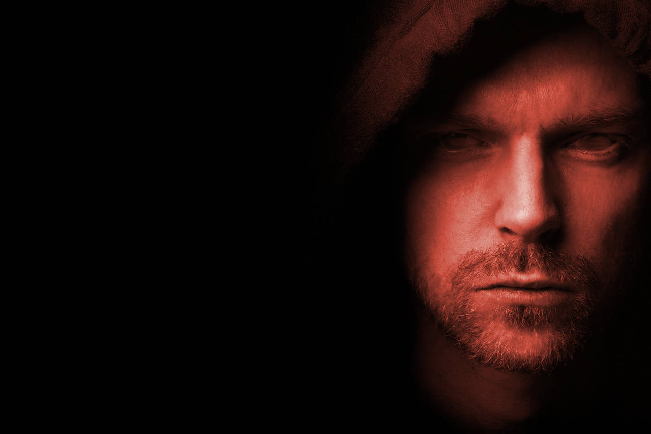 A lightly red tinted photo of the headshot of a Caucasian man with a mustache and light beard positioned to the far right wearing a brown hood and displayed with a serious expression on his face