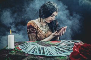 A Caucasian female fortune-teller with long black hair and a light brown dress seated in front of a table filled with an array of tarot cards, a candle, and a red rose while looking down at one of the cards held up in her hands