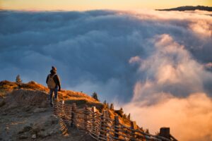 A man dressed in winter outerwear wearing a backpack standing in the top of a mountain overlooking a vast valley filled with clouds with a setting orange winter sun streaming across the area