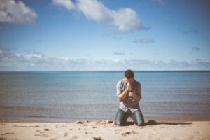 A man with black hair, light grey shirt, and blue jeans kneeling with his head bowed, hands raised and clasped together on a beach in front of a large calm body of water and a lightly clouded blue sky in the background