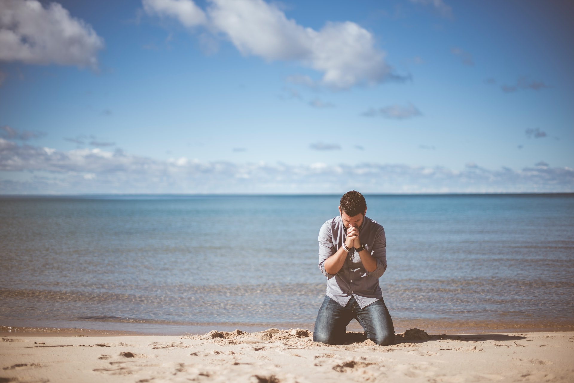 A man with black hair, light grey shirt, and blue jeans kneeling with his head bowed, hands raised and clasped together on a beach in front of a large calm body of water and a lightly clouded blue sky in the background