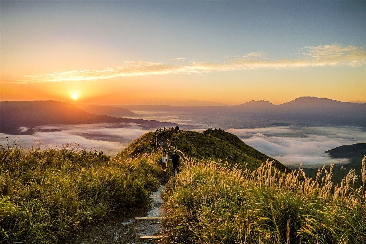 A small path on a narrow grassy hillside leading towards a fog laden valley in the midst of several large mountains with the sun rising on the left in the distance and a pale blue sky with a small band of clouds overhead