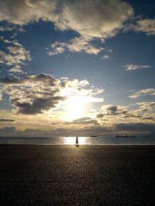 A setting sun, partially obscured behind clouds in a blue sky over a large body of water, highlighting a man walking on the sandy shoreline