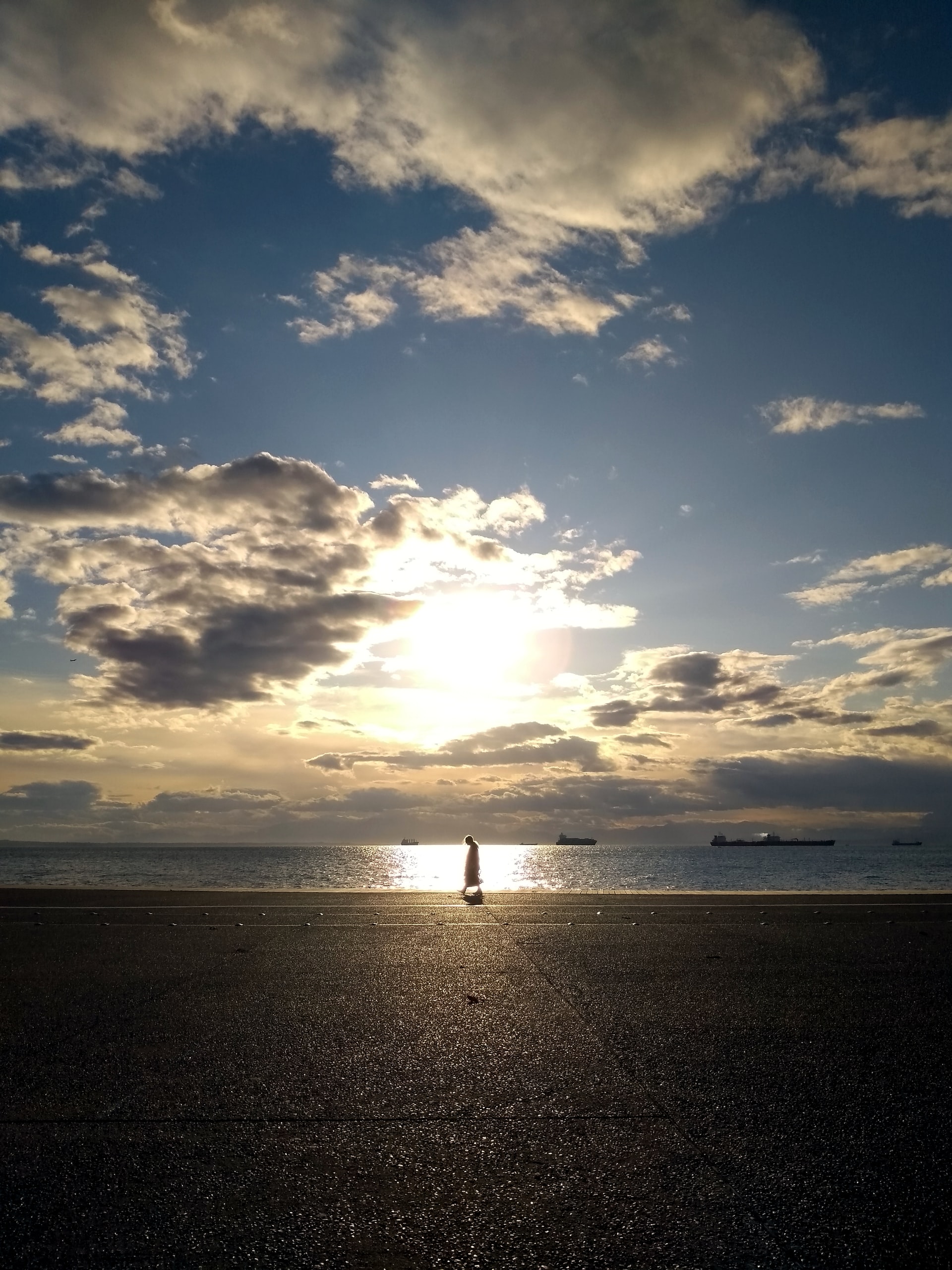 A setting sun, partially obscured behind clouds in a blue sky over a large body of water, highlighting a man walking on the sandy shoreline