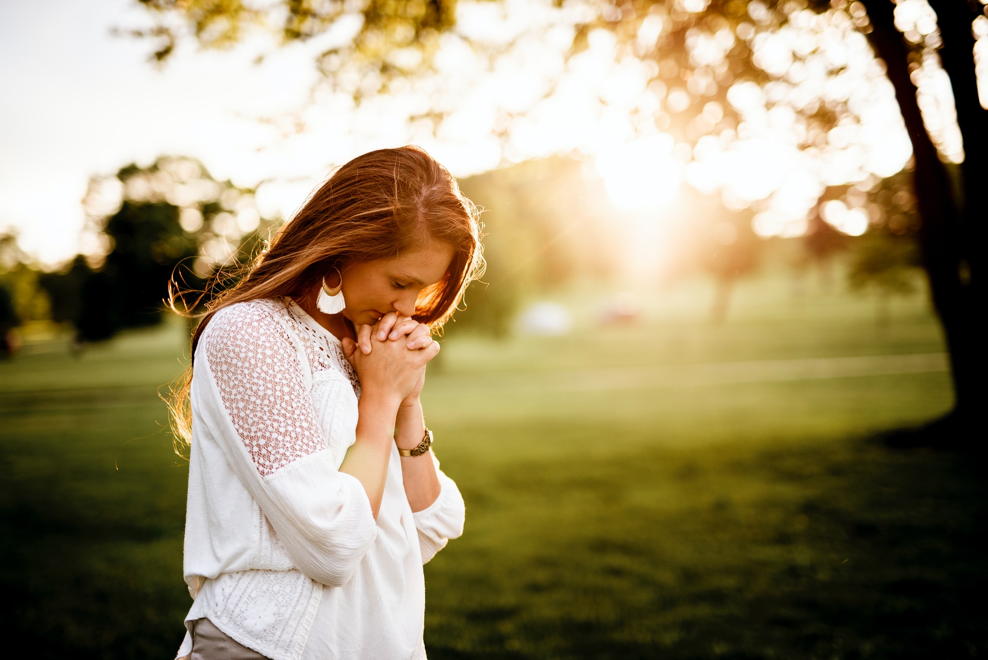 A photo of a Caucasian woman with long brown hair and a white blouse standing in a field with trees while bowing her head against her folded hands in prayer