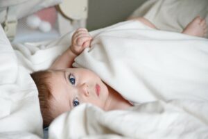 A photo of a baby with brown hair and blue eyes lying in a bed between white covers looking towards the viewer with small crib in the background