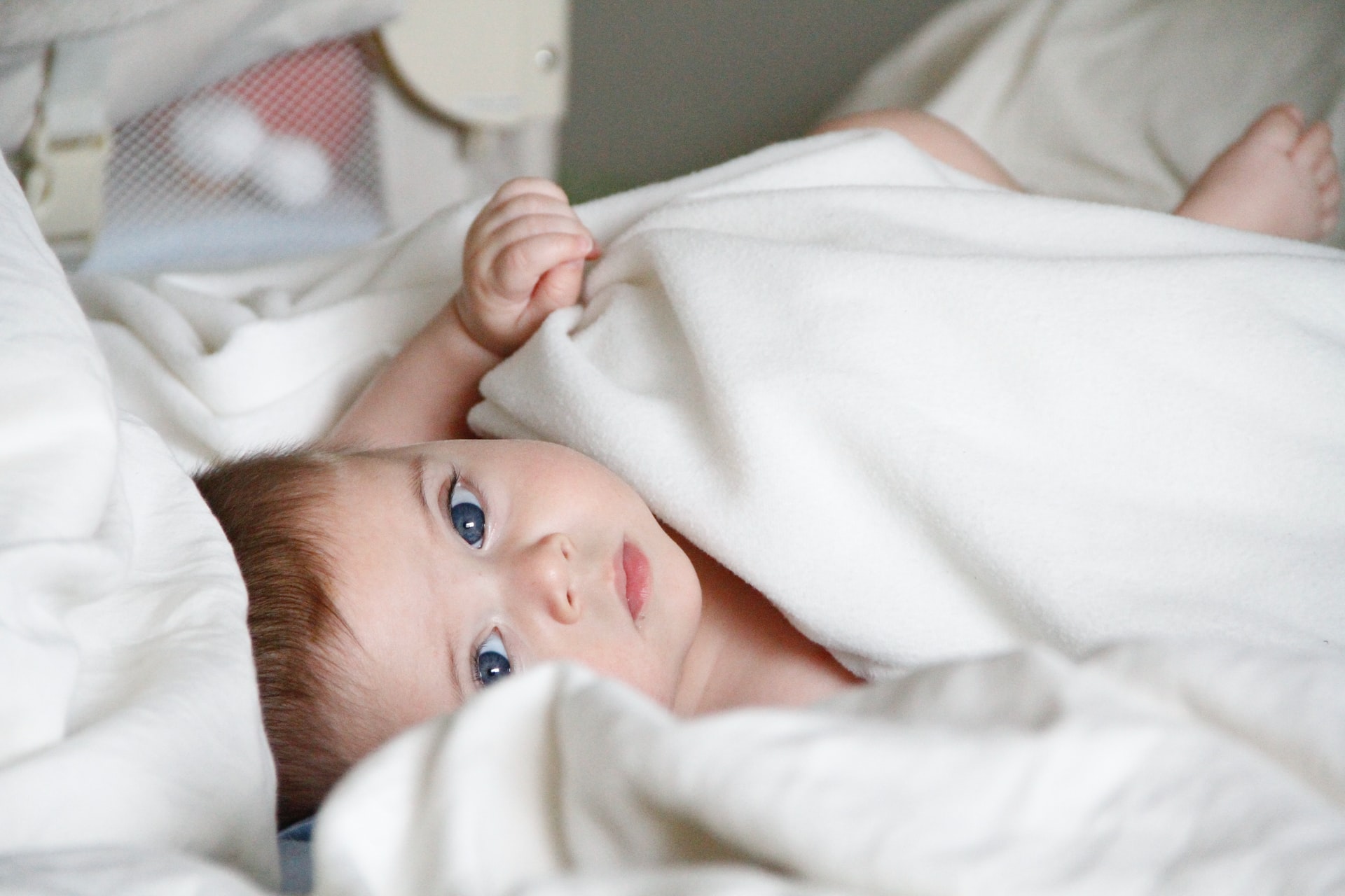 A photo of a baby with brown hair and blue eyes lying in a bed between white covers looking towards the viewer with small crib in the background