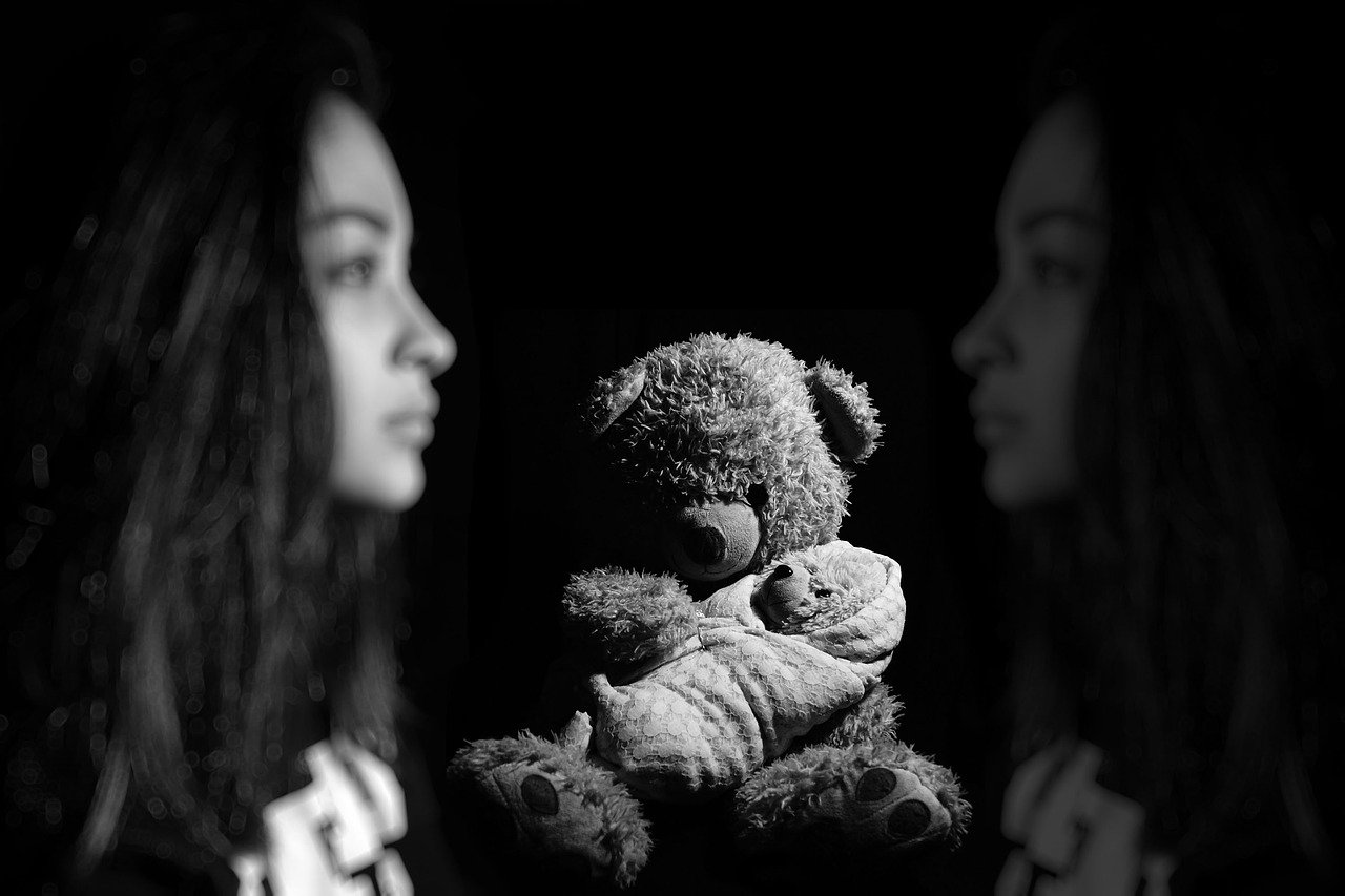 A black and white photo of a small stuffed bear located front and center between a young African-American woman looking to the right and a muted reflection of herself looking left