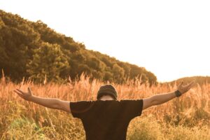 A photo of the upper backside of a young Caucasian man wearing a black t-shirt and a grey knit hat with his head leaning downward and arms outstretched against a field of tall dry grasses and a line of trees in the background