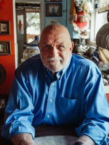 An older bald Caucasian man seated in a well lit room wearing a medium blue long sleeved shirt with various pictures and household objects around him