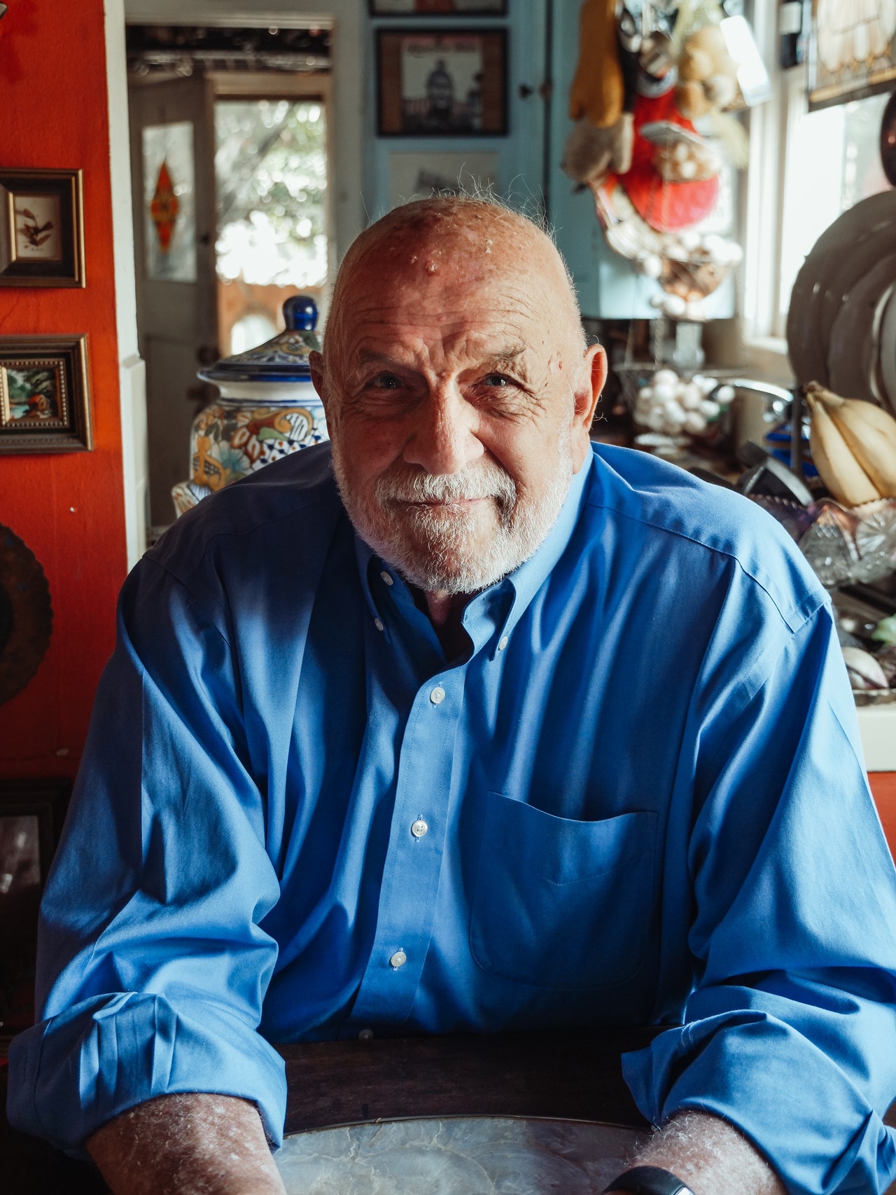 An older bald Caucasian man seated in a well lit room wearing a medium blue long sleeved shirt with various pictures and household objects around him