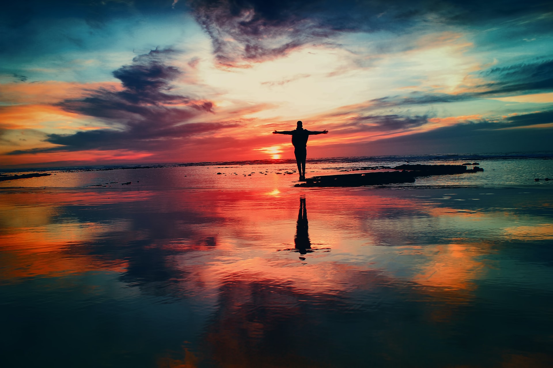 A silhouette of a man standing on a small cluster of rocks in the midst of a large body of water with his arms outstretched and a multicolored sunset in the background directly reflecting on the water