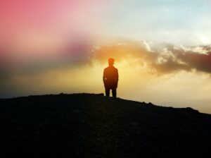 A photo of the silhouette of the back of a man standing on a small hill looking out towards a cluster of white clouds with an iridescent haze over most of the scene