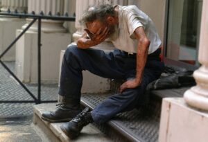 An older Caucasian man with his right hand clutched over his face and his left supported on his left knee seated on a small set of steps of a building with large white columns