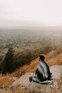 A young Caucasian man with dark hair and glasses seated on a rock of a small mountain looking out over the foggy forest in the valley