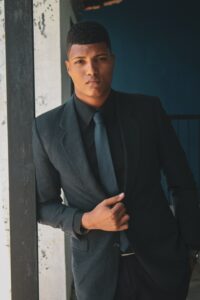 A light skinned African-American man wearing a dark suit with a dark blue tie and leaning against a column in an old building