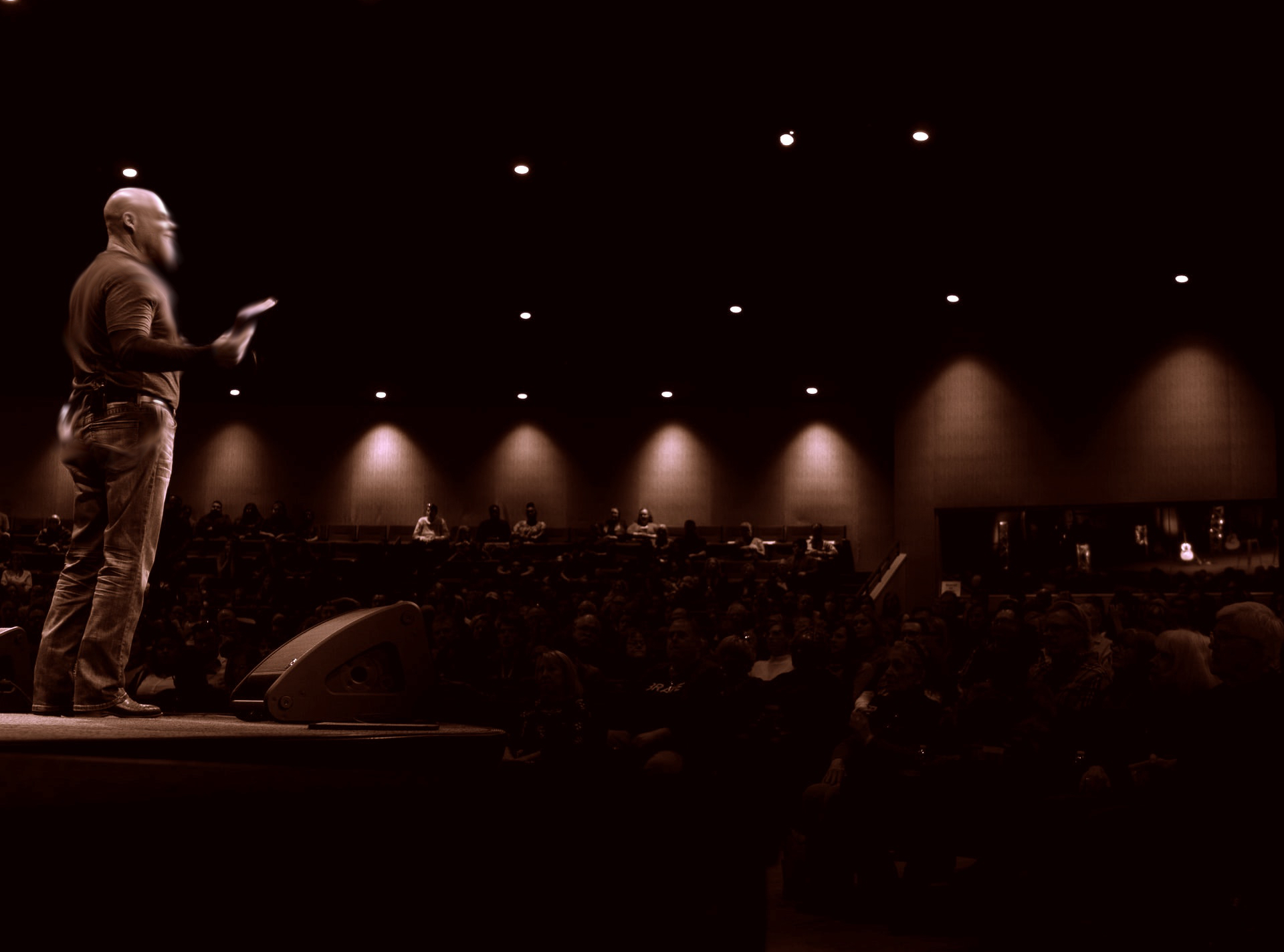 A minister in a long sleeved shirt and jeans holding a Bible while speaking to a large crowd in a mostly darkened room symbolizing religious deception by Christian leaders