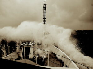 A tinted black and white photo looking across the bow of a ship with a large wave crashing over it and a clouded sky in the background