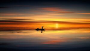 A small boat with a person sitting and another standing while holding a fishing rod in the midst of a vast calm body of water mirroring in its reflection a golden setting sun among thin clouds