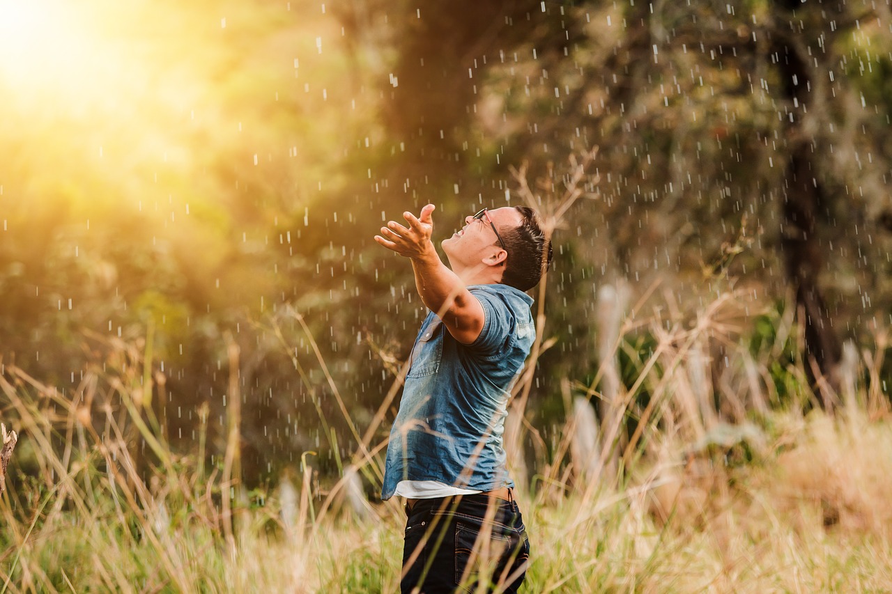 A photo of a man wearing a blue short sleeved shirt and black pants standing in a light rain in a field of tall grasses with trees and sunlight in the background holding his hands up and looking upward in worship