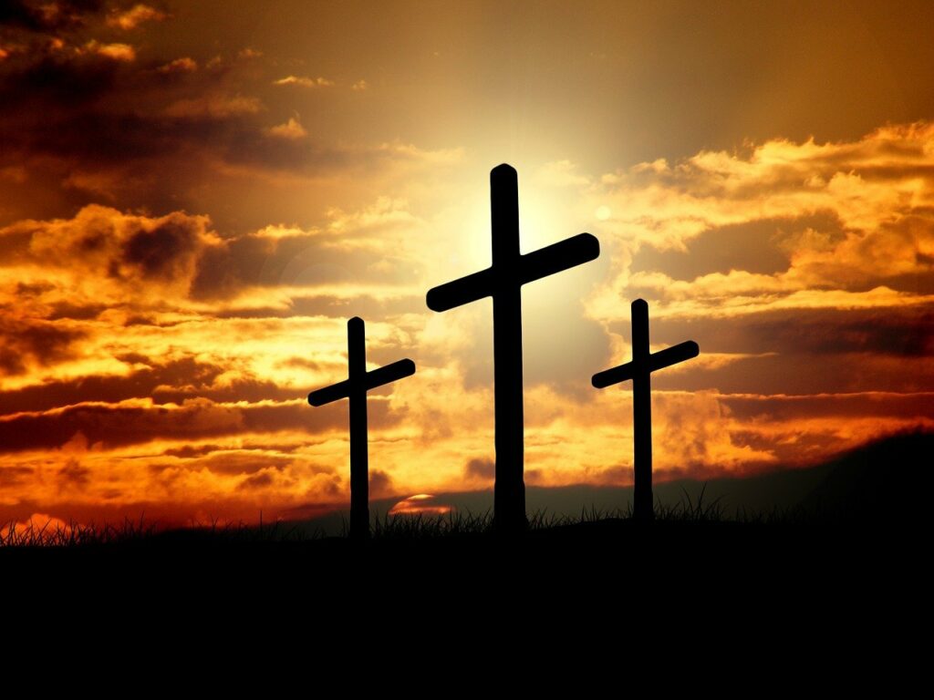 A silhouette of three crosses with an orange illuminated clouded sky in the background and the sun glowing behind the center cross