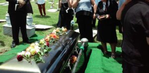 A photo of the waist shot of several people standing around a flower strewn casket in a funeral on a bright sunny day