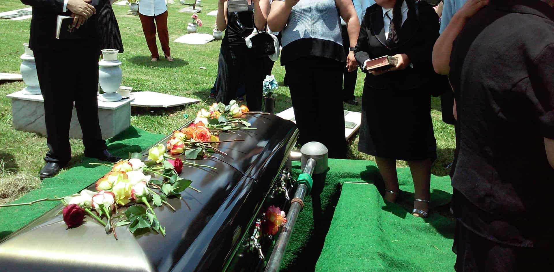 A photo of the waist shot of several people standing around a flower strewn casket in a funeral on a bright sunny day