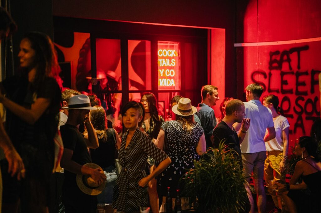 A mixed group of people standing around in a cocktail bar with red walls with words spray painted in black on one wall and an orange neon sign in the main window with words 'cocktails y coviida'