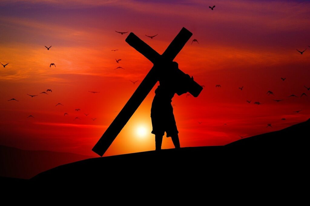 A silhouette of a man carrying a large cross up a small hill with a red sky, a low setting yellow sun, and many flying birds in the background