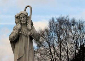 A photo of a sculpture of Jesus holding a shepherd's staff and a lamb over the back of His neck with a tree silhouette and a partially cloudy blue sky in the background
