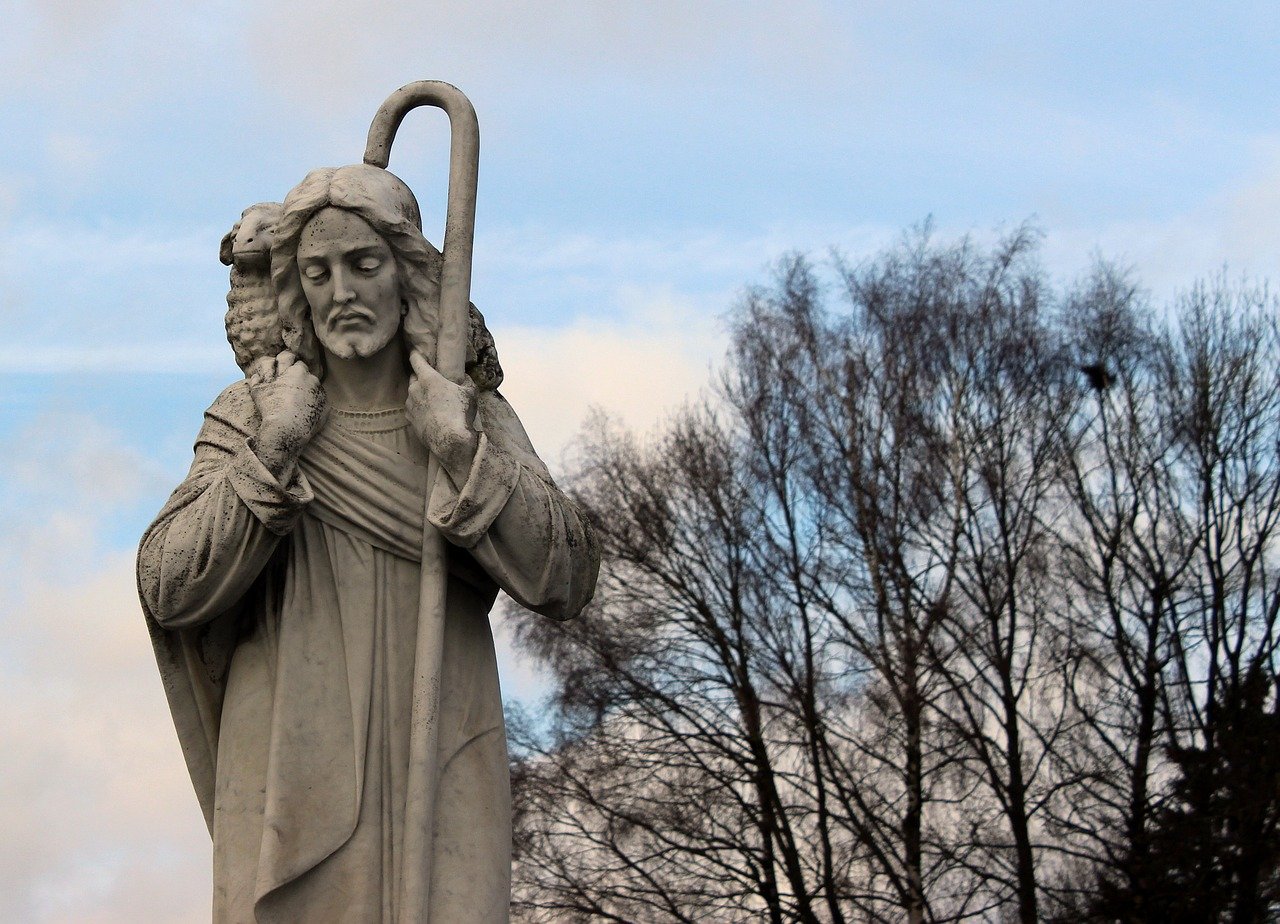 A photo of a sculpture of Jesus holding a shepherd's staff and a lamb over the back of His neck with a tree silhouette and a partially cloudy blue sky in the background