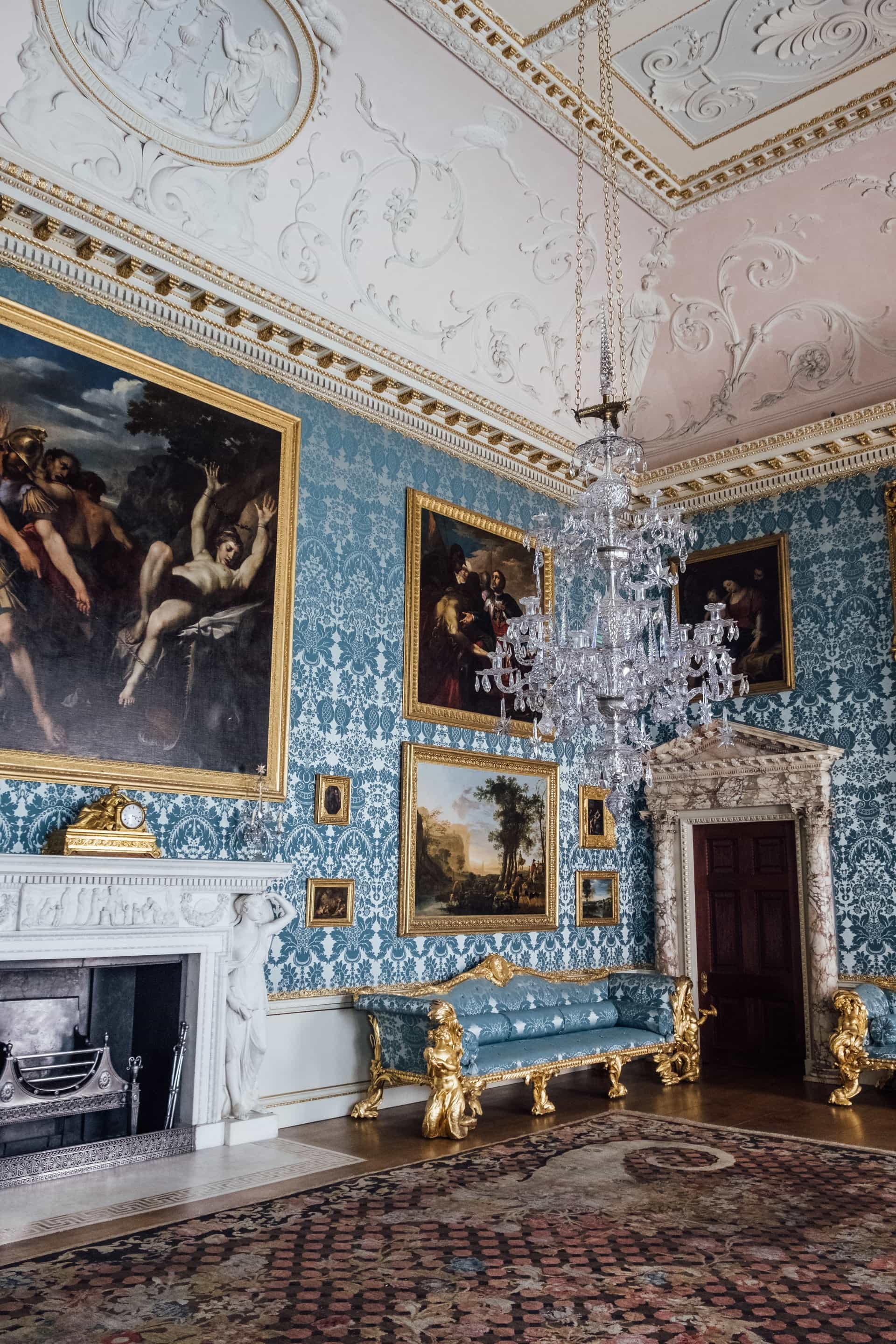 A lavish Victorian style room with many ornate paintings on the walls, blue print wallpaper with several types of exquisite moldings, a large glass chandelier, a near wall to wall Oriental rug, fireplace, gold trimmed furniture upholstered with the same pattern as the wallpaper all placed in a very high ceiling room