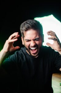 A Caucasian young man with black hair wearing a black t-shirt while holding both of his hands up toward his head and expressing a loud scream with his eyes closed