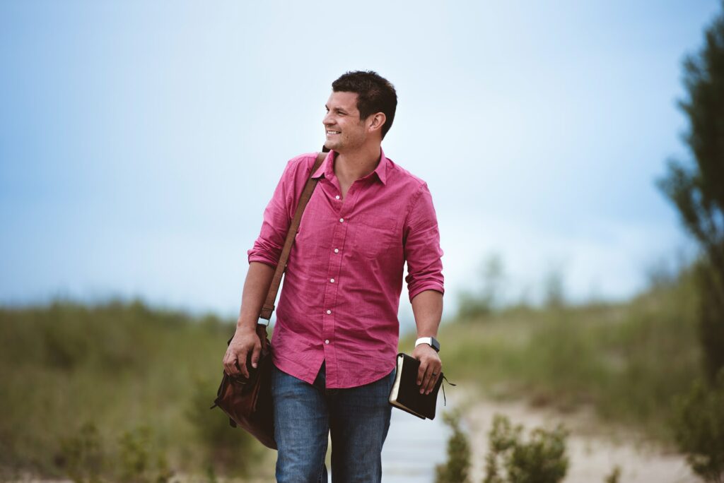 A photo of a Caucasian man with black hair, magenta buttoned shirt, and blue jeans walking down a path near a seashore while smiling, carrying a leather bag over his right shoulder, and holding a Bible in his left hand