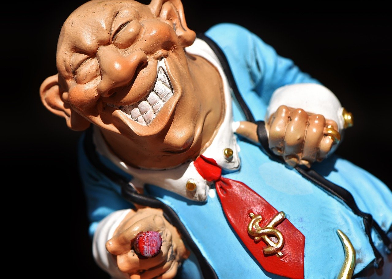A photo of a toy character modeled after a bald headed, chubby, smiling loan shark wearing a blue shirt with white cuffs, a red tie with a gold dollar sign, and a large gold horned cattle head belt buckle while holding his suspenders and pointing at the camera with a red tipped cigar