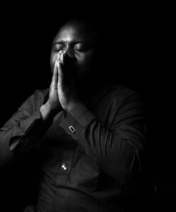 A black and white photo of a Nigerian man in a black shirt with eyes closed and holding his hands pressed together up to his face in a position of prayer