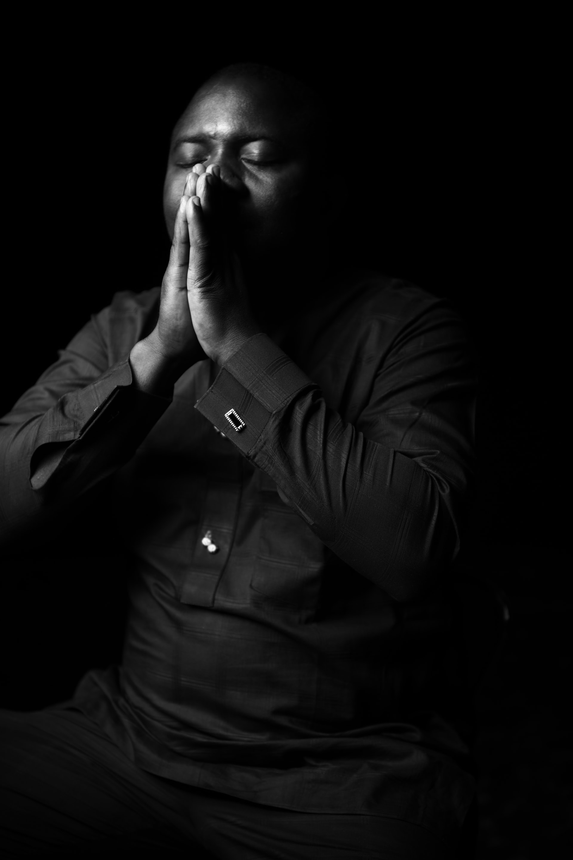 A black and white photo of a Nigerian man in a black shirt with eyes closed and holding his hands pressed together up to his face in a position of prayer