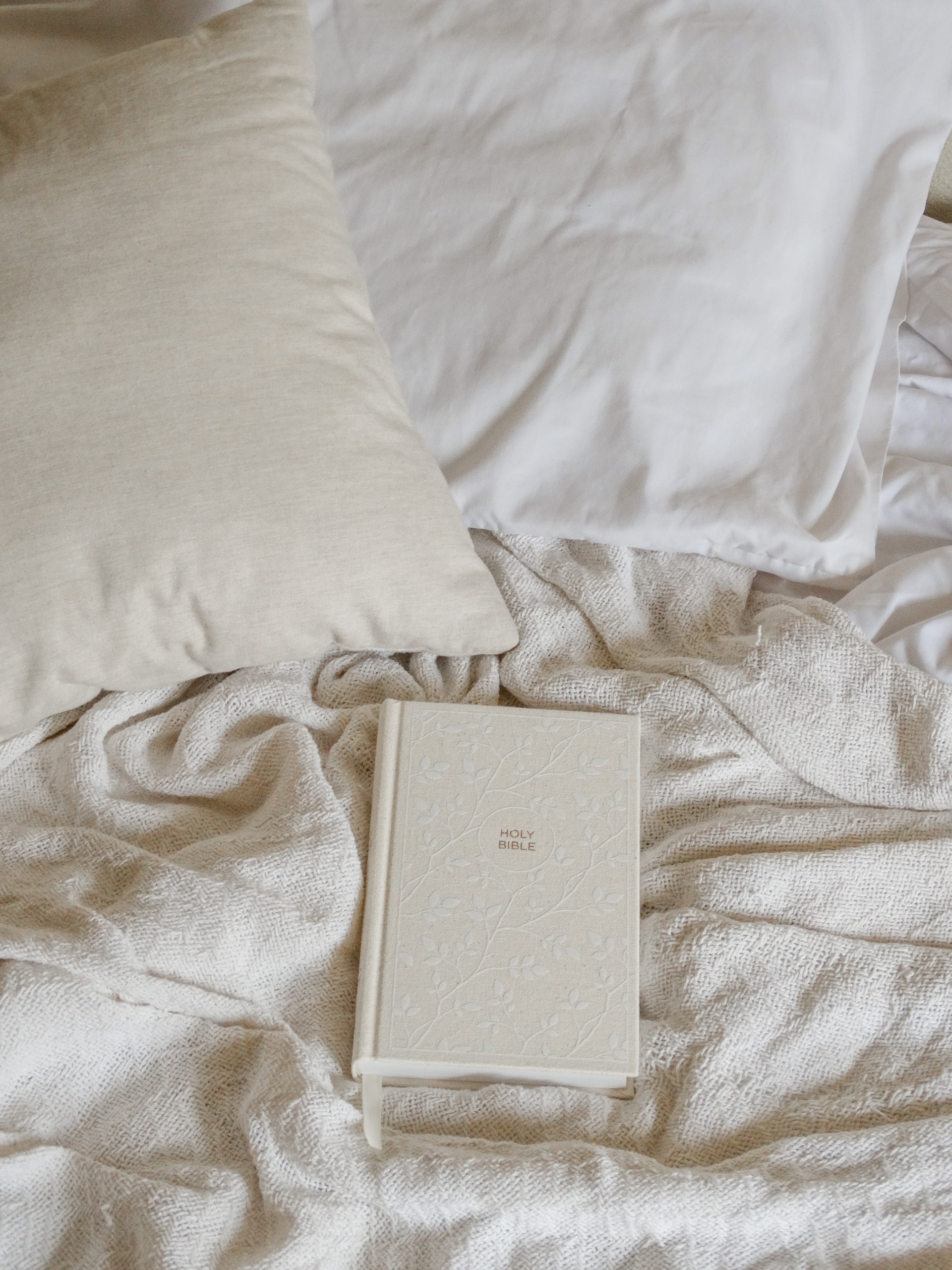 A photo of an off-white Bible laying in the midst of a bed covered with white and off-white sheets and pillows