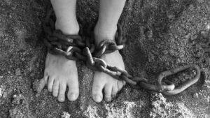 A black and white photo of a pair of bare feet resting in sand wrapped in a section of old heavyweight chain